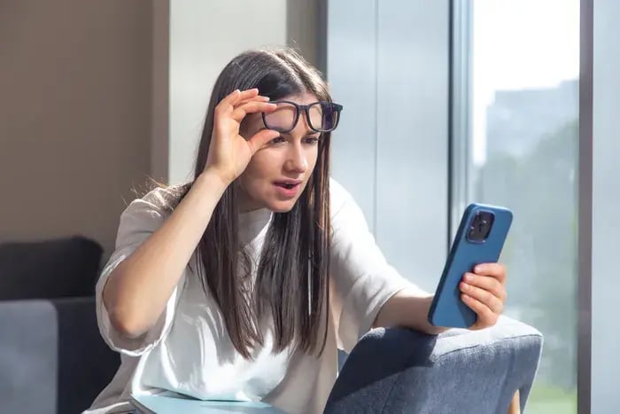 Woman lifting her glasses while looking at a smartphone by the window.